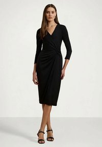 Mulher vestindo um vestido envolvente preto até ao joelho com mangas três quartos e tacões abertos pretos, de pé contra um fundo simples.