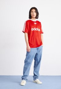 Rode Adidas sportshirt met witte accenten en logo, gecombineerd met lichtblauwe jeans en crèmekleurige sneakers. Soepele textuur, relaxte pasvorm, korte mouwen.