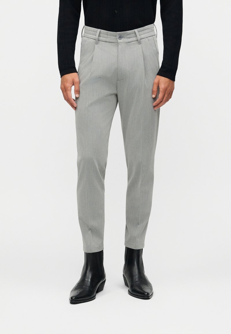 Hombre vistiendo pantalones personalizados de color gris claro, camiseta negra de manga larga y botines de cuero negros con punta, de pie contra un fondo blanco.