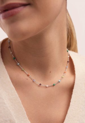 Primer plano del cuello de una persona que lleva un delicado collar de cadena plateada con pequeñas cuentas multicolores distribuidas uniformemente a lo largo de él.