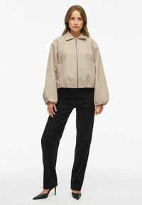 Bežs īsais žakets ar rāvējslēdzēju, paceltu apkakli un elastīgām aprocēm, saskaņots ar melnām taisnas kājas biksēm un melniem smailiem papēžiem.