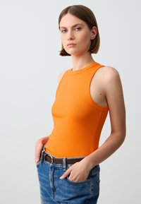Top arancione senza maniche dal design aderente, realizzato in cotone elasticizzato, abbinato a jeans in denim blu a vita alta e una cintura in pelle marrone.