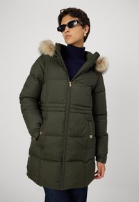 Olīvzaļš pildīts mētelis ar mākslīgās kažokādas kapuci, ar rāvējslēdzēju, sānu kabatām un zeltainiem aksesuāriem. Tekstūrēta virsma ar šuvēm.