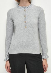 Pull en laine grise clair avec un col volanté, une patte de boutonnage à cinq boutons et des poignets volantés. Texture douce et design ajusté.