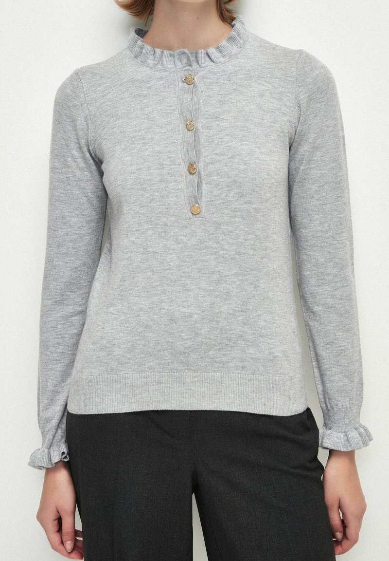 Pull en laine grise clair avec un col volanté, une patte de boutonnage à cinq boutons et des poignets volantés. Texture douce et design ajusté.