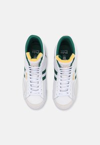 Sneakers bianchi con accenti verdi e gialli. Le caratteristiche includono pelle liscia, pannelli in rete e un branding prominente sulla linguetta e sui lati.