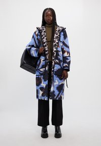 Κουβερτίνα σε μπλε, μαύρο και ροζ λουλουδάτο σχέδιο με κουκούλα και μαύρες λεπτομέρειες. Συνδυάζεται με μια ματ μαύρη τσάντα και μαύρα παντελόνια.