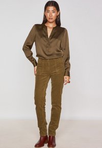 Blusa di raso verde oliva con scollo a V e maniche lunghe, abbinata a pantaloni in corduroy beige con tasche. Il modello indossa stivali alla caviglia marroni.