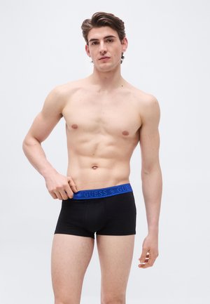 Νεαρός άνδρας με κοντά καστανά μαλλιά που φοράει μαύρα μπόξερ με μπλε ζώνη μέσης "GUESS", ρυθμίζοντας τη ζώνη μέσης με το ένα χέρι.