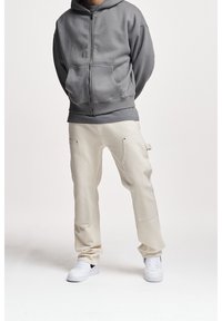 Grijze rits hoodie met voorzakken, gecombineerd met beige cargo broeken met zijzakken en witte sneakers. Effen achtergrond.