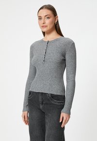 Grijze geribbelde gebreide trui met een ronde hals en knoopdetails, met lange mouwen en een getailleerde taille. Gepaard met een donkere spijkerbroek.