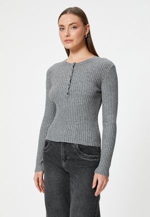 Grijze geribbelde gebreide trui met een ronde hals en knoopdetails, met lange mouwen en een getailleerde taille. Gepaard met een donkere spijkerbroek.