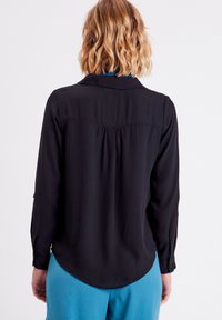 Blouse noire à manches longues avec un col, en tissu texturé et un ourlet arrondi. Dispose d'une couture de carrure au dos et de poignets à boutons.