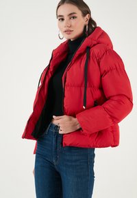 Rød jakke i puffer-stil med hætte, der har sorte snore, polstret design, lynlåslommer i siderne og en cropped længde over mørke jeans.