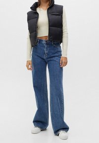 Black puffer vest with high collar, beige long-sleeve top, high-waisted flared blue jeans, and white sneakers on a light background.