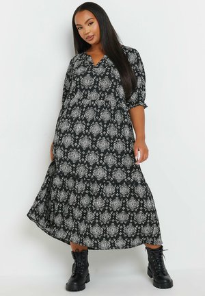 Zwarte jurk met witte ornamentale patronen, v-hals, driekwart mouwen, vloeiende silhouet, gecombineerd met zwarte veterschoenen.