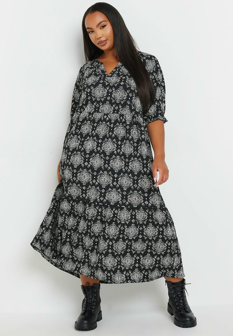 Musta mekko, jossa on valkoisia koristeellisia kuvioita, v-pääntie, kolmen neljänneksen hihat, aaltoileva siluetti, yhdistettynä mustiin nauhakenkiin.