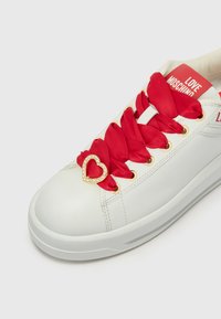 Weiße Leder-Sneaker mit roten Satinschnürsenkeln, goldenen Ösen und einer herzförmigen Strassdekoration an den Schnürsenkeln. "LOVE MOSCHINO" auf der Zunge gedruckt.