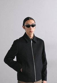 Giacca nera con colletto e zipper frontale, abbinata a occhiali da sole a forma di occhio di gatto neri con dettagli borchiati. Texture liscia, design minimale.