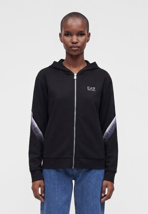 Persoon draagt een zwarte hoodie met rits met EA7 Emporio Armani-logo's op borst en mouwen, gecombineerd met een blauwe spijkerbroek, staand tegen een effen achtergrond.