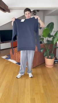 Tamsiai mėlynas džemperis su priekine kišene, ilgomis rankovėmis ir su raišteliu reguliuojama kapuce, eksponuojamas šviesioje patalpoje su žaluma.
