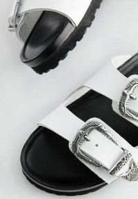 Vita sandaler med två justerbara remmar, försedd med ornamenterade silverfästen, en texturerad svart sula och en slät materialyta.