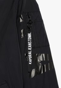 Musta nailonitakki kiiltävillä mustilla yksityiskohdilla, jossa on sivuvetoketjullinen tasku ja selkeä brändilogo painettuna pystysuoraan hihaan.