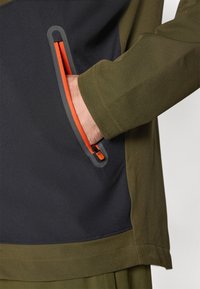 Alyvuogių žalia striukė su tamsiai mėlynu intarpų, turinti oranžinį užtrauktuko kišenę ir elegantišką, tekstūruotą audinio apdailą. Tvarkingas, modernus dizainas.