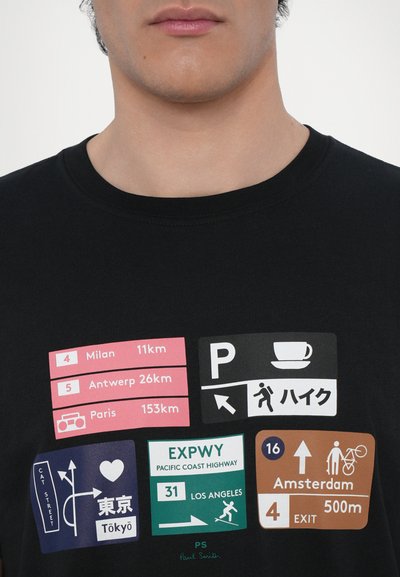 T-shirt noir avec des graphismes colorés de panneaux de signalisation et des distances vers diverses villes. Comprend des icônes pour le stationnement et la marche.