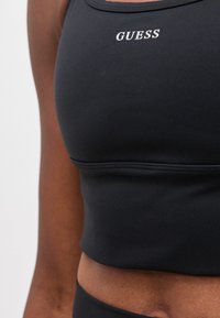 Κοντινό πλάνο ενός ατόμου που φοράει ένα μαύρο αθλητικό σουτιέν με το λογότυπο "GUESS" κεντημένο με άσπρο στη θώρακα.