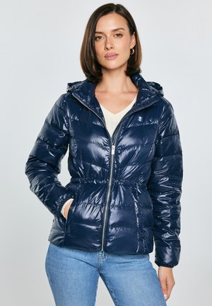 Naine, kes kannab läikivat tumelilla tuhvlijopet kapuutsiga, esimesel lukuga, valge kampsuni ja hele siniste teksadega, seisab ühe käega taskus.