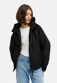 Svart huvjacka med dragkedja framtill, med en lös passform, elastiska mudd och dragsko-detaljer. Bärs över en vit skjorta och jeans.