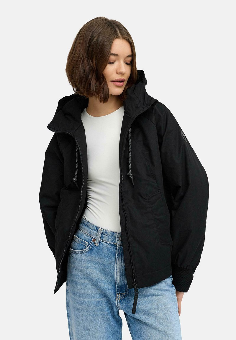 Svart huvjacka med dragkedja framtill, med en lös passform, elastiska mudd och dragsko-detaljer. Bärs över en vit skjorta och jeans.