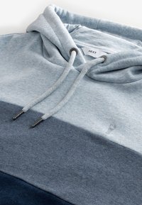 Sweat à capuche avec des rayures horizontales bleu clair, moyen et foncé, avec cordons et une étiquette de marque « NEXT » à l’intérieur du col.