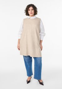 Béžová pletená bezrukávová tunika s okrúhlym výstrihom, skombinovaná s voľnými modrými džínsovými nohavicami a čiernymi špicatými podpätkami. Pod tunikou biela košeľa.