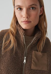 Veste zippée en polaire marron avec une surface texturée et une poche en tissu beige contrastante. Dotée d'un col haut et d'une fermeture éclair métallique.