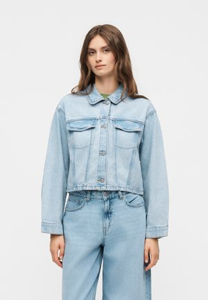 Ung kvinde iført lyseblå denimjakke og matchende jeans, stående mod en ensfarvet hvid baggrund, vendt mod kameraet.