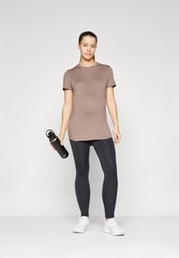 Brun kortermet trening t-skjorte, elastiske svarte leggings, hvite joggesko, holder en svart vannflaske med lokk, ensfarget bakgrunn.