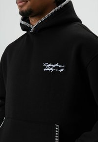 Zwarte hoodie met witte decoratieve stiksels op de capuchon en zak. Voorzien van een geborduurd tekstlogo op de borst. Zachte stoftextuur.