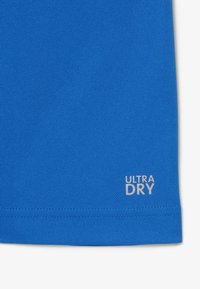 Gros plan sur un tissu bleu avec "ULTRA DRY" imprimé en blanc près du bord inférieur, montrant un tricot texturé et une couture d'ourlet.