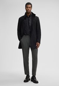 Uomo che indossa un cappotto nero a strati, una camicia nera, pantaloni grigi strutturati e scarpe eleganti nere, in piedi con le mani in tasca contro uno sfondo semplice.