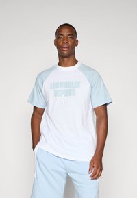 Hvit t-skjorte med lyseblå raglan-ermer, med "LOS ANGELES DODGERS" trykket i lyseblått. Kombinert med matchende lyseblå shorts.