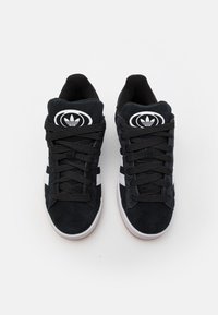 Chaussures de sport en suede noir avec des rayures et un logo blancs. Elles sont dotées d'une languette rembourrée, de lacets épais et d'une semelle en caoutchouc gomme texturée.