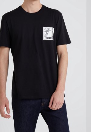 Mann trägt schwarzes Versace-T-Shirt mit weißem Logopatch auf der linken Brust und dunkelblaue Jeans, steht vor weißem Hintergrund.