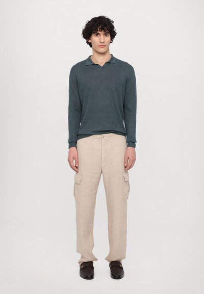 Donkergroene polo met lange mouwen en kraag, gecombineerd met lichtbeige cargobroeken met zijzakken en een gladde textuur. Bruine schoenen.