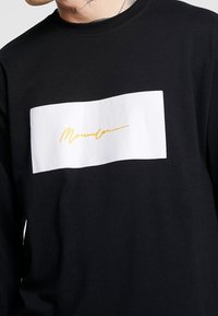 Schwarzes Langarm-T-Shirt aus Baumwolle mit einem weißen rechteckigen Aufnäher und einem gelben Signaturdesign auf der Brust.