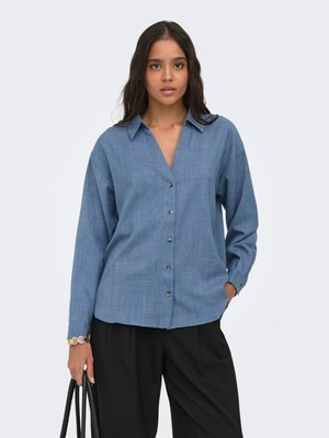 Frau trägt ein blaues Hemd mit Knöpfen und schwarze Hose, hält eine schwarze Tasche und steht vor einem schlichten weißen Hintergrund.