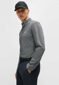 Grå skjorta med knappar och ett strukturerat mönster, med krage, långa ärmar och en liten vit logotyp på vänster sida. Svart keps och bälte.