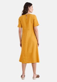Hořčicově žluté lněné šaty s krátkými rukávy, ke kolenům, s výřezem na zádech a s mírným A-liniovým tvarem. Hladká textura a jednoduchý design.
