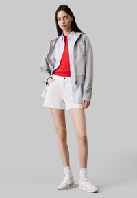 Sudadera gris con cremallera, camiseta roja y camisa de botones azul clara superpuestas, combinadas con pantalones cortos blancos deshilachados y zapatillas blancas.